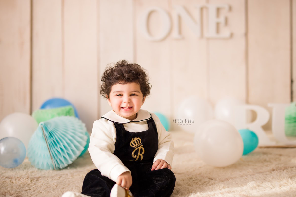 Immerse in the delightful innocence of a 1-year-old girl amidst a serene scene: white and blue balloons against a soothing beige backdrop, complemented by a playful pompom beige rug. This charming moment is captured through a sitter photoshoot in Delhi and Gurgaon by Anega Bawa.