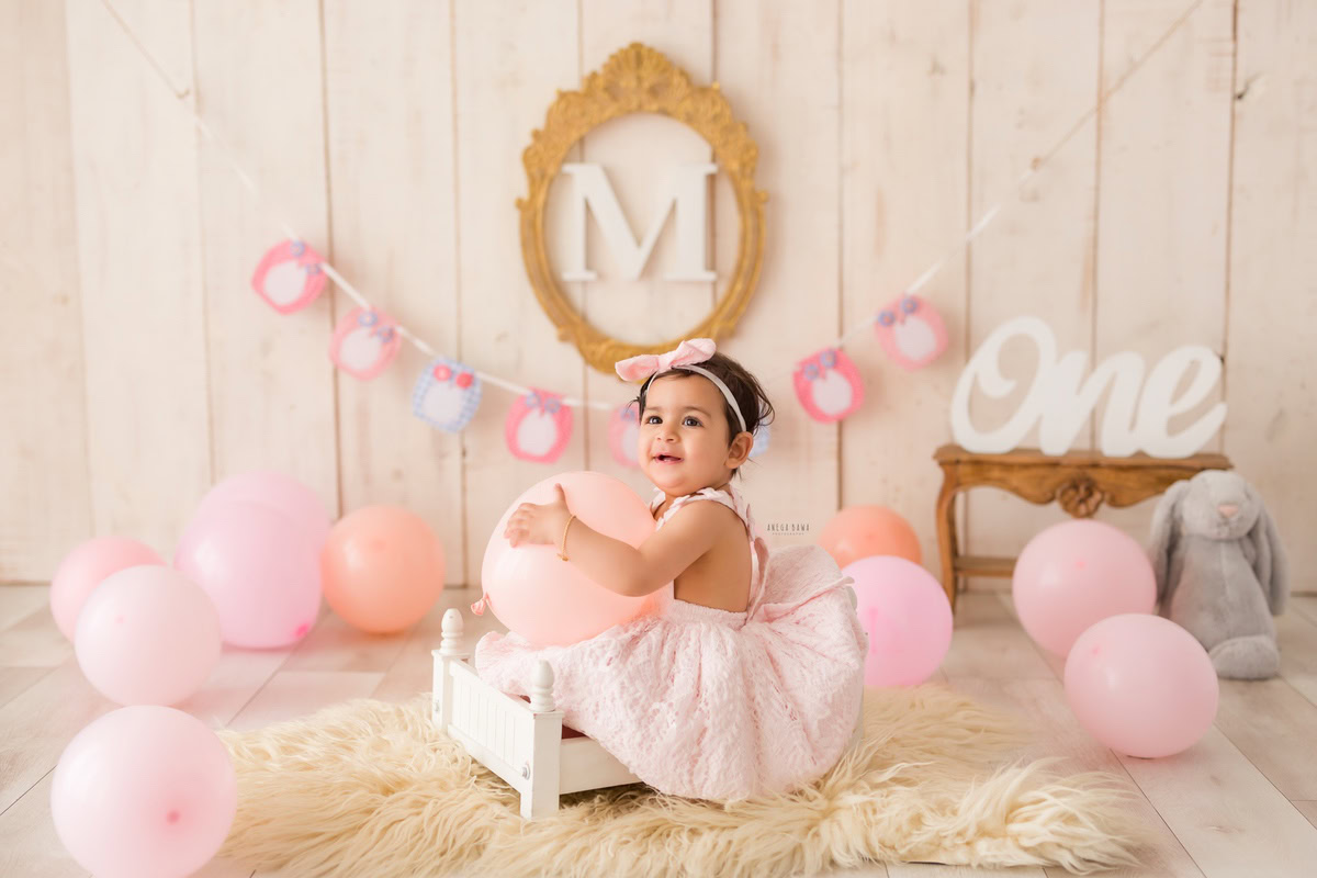 Witness the delightful scene of a 1-year-old girl seated in a white cot, amidst a backdrop adorned with peach-pink balloons and a wooden frame on the wall. This captivating moment was expertly captured during a toddler photoshoot in Delhi, Gurgaon, and Noida by the talented photographer, Anega Bawa.
