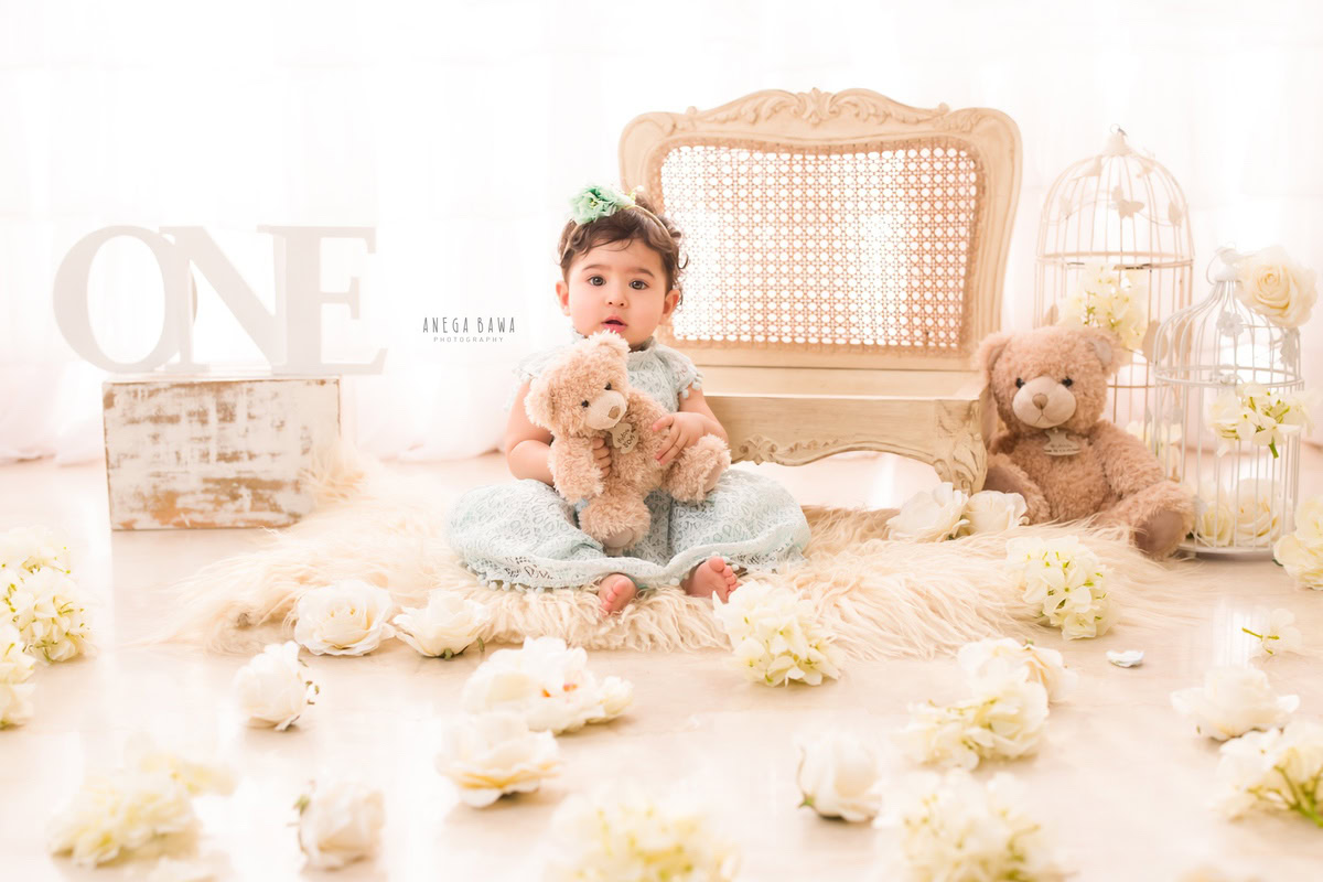 Delight in the innocence of your 1-year-old girl's sitter photoshoot captured by Anega Bawa Photography in Delhi and Gurgaon. Adorned with white flowers and surrounded by wooden chairs and teddy bears against a pristine white backdrop, each snapshot preserves the magic of her early years.