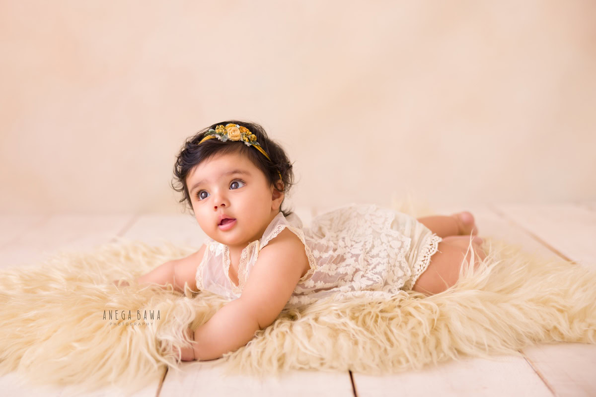 Charming 12-month-old girl lying on a cozy beige rug against a soft beige backdrop, wearing a cute headband, in her first birthday photoshoot in Delhi by Anega Bawa, Gurgaon, Noida
