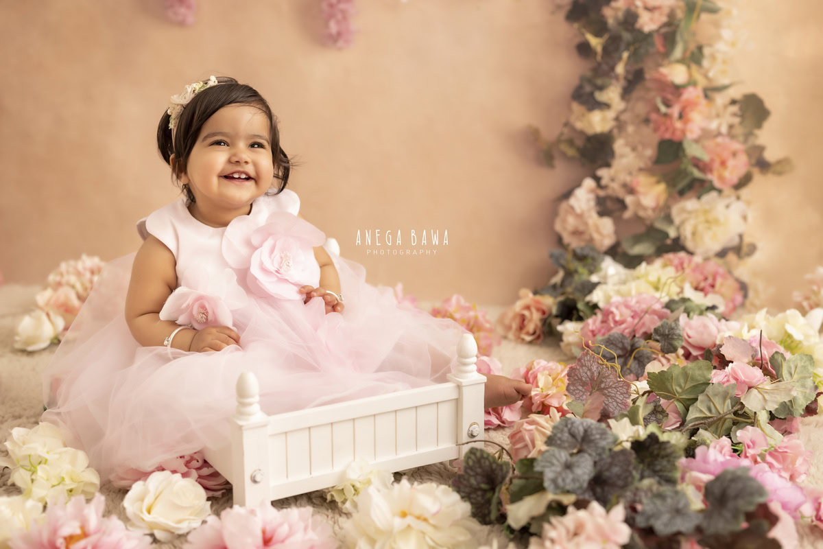 Witness the adorable scene of a 15-month-old girl seated in a white cot, adorned in a charming pink dress, surrounded by a plethora of multicolored flowers. This delightful moment was expertly captured during a toddler photoshoot in Delhi, Gurgaon, and Noida by the talented photographer, Anega Bawa.