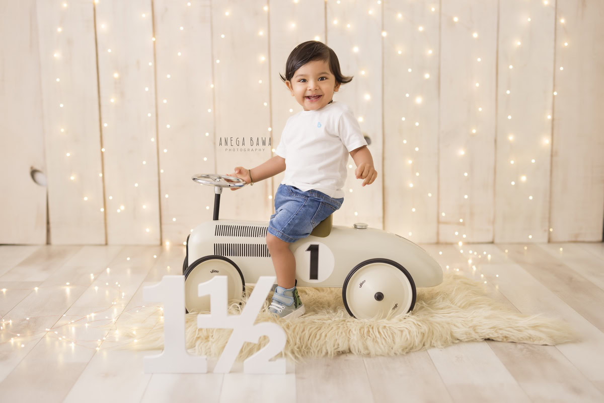 In this delightful capture from a baby pre-birthday photoshoot in Delhi by Anega Bawa, an adorable 18-month-old boy sits confidently on a toy scooter, surrounded by the soft glow of fairy lights. The beige rug beneath him provides a cozy setting for his playful adventure. This charming scene captures the innocence and joy of childhood, creating a cherished memory for years to come.