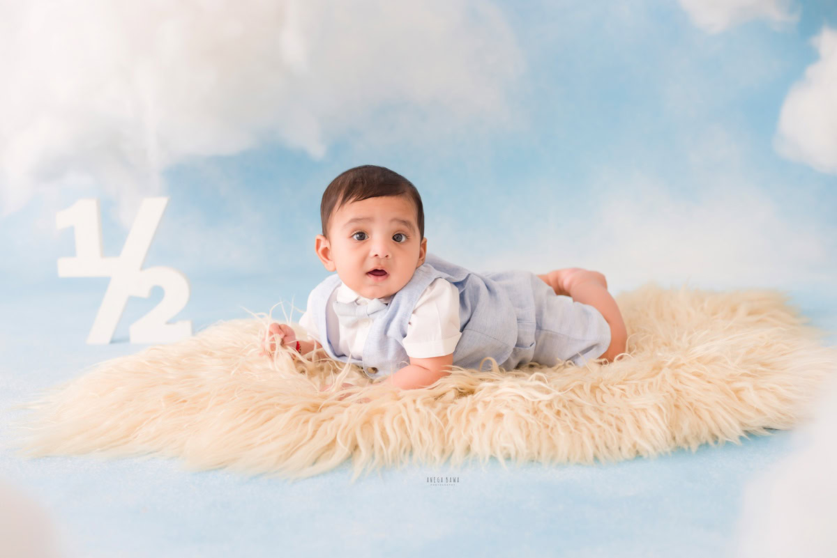 In this captivating image from a baby pre-birthday photoshoot in Delhi by Anega Bawa, a 6-month-old boy is nestled on a plush beige rug, set against a dreamy cloudy backdrop. The soft lighting and tranquil atmosphere perfectly capture the innocence and wonder of infancy, creating a cherished memory to treasure for years to come.