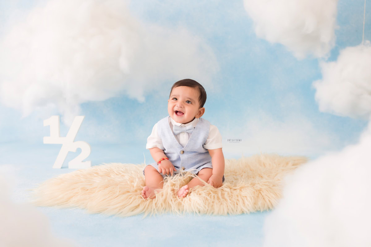 In this enchanting capture from a baby pre-birthday photoshoot in Delhi by Anega Bawa, a joyful 6-month-old boy beams with pure delight as he rests on a cozy beige rug. The whimsical cloudy backdrop adds a touch of magic to the scene, enhancing the innocence and charm of the moment. It's a heartwarming image that beautifully encapsulates the joy and wonder of infancy, creating a treasured memory for years to come.