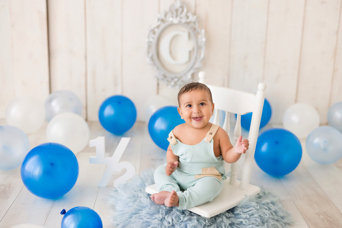 Marvel at the precious sight of a 6-month-old boy seated on a white chair, surrounded by a blue rug and delicate blue and white balloons against a beige backdrop. This charming moment was expertly captured during a toddler photoshoot in Delhi, Gurgaon, and Noida by the skilled photographer, Anega Bawa.