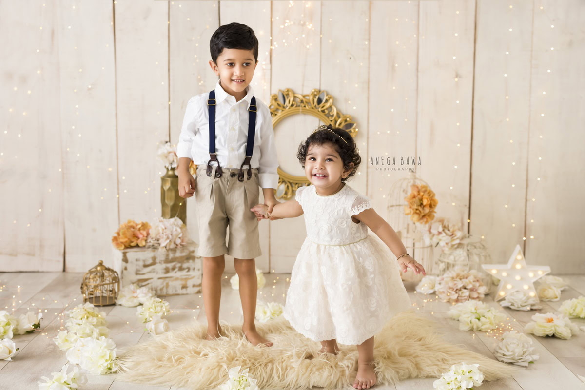 For a baby pre-birthday photoshoot in Delhi, Anega Bawa, Gurgaon, and Noida, siblings sit together on a beige rug against a beige backdrop adorned with castles and fairy lights. This serene and enchanting scene captures the bond between the siblings as they celebrate this special occasion together, surrounded by magical elements that add a touch of whimsy to the moment.