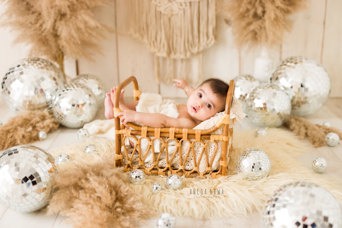 Capture the innocence and joy of your 3, 4, 5, or 6-month-old baby boy wrapped in beige, nestled in a wooden cot, surrounded by twinkling disco balls on a cozy beige rug against a neutral backdrop. Contact Anega Bawa for your baby boy's photoshoot in Delhi, Gurgaon, or Noida.