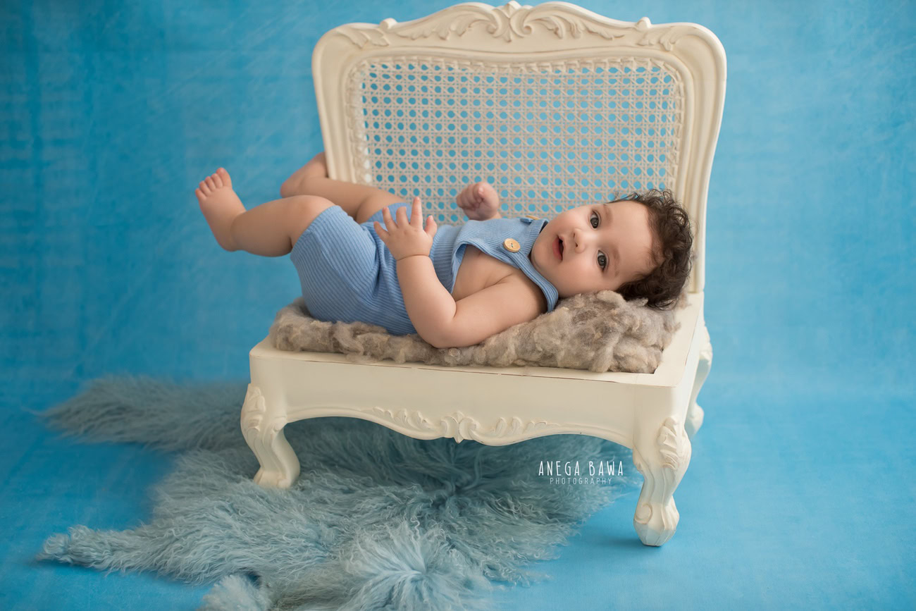 Mesmerizing baby boy photo shoot, showcasing precious 6, 5, 3, 4-month-old moments against a serene blue backdrop, accented with a cozy grey rug and a pristine white wooden chair. Expertly composed by Anega Bawa, the premier baby photographer in Delhi, Gurgaon, and Noida.