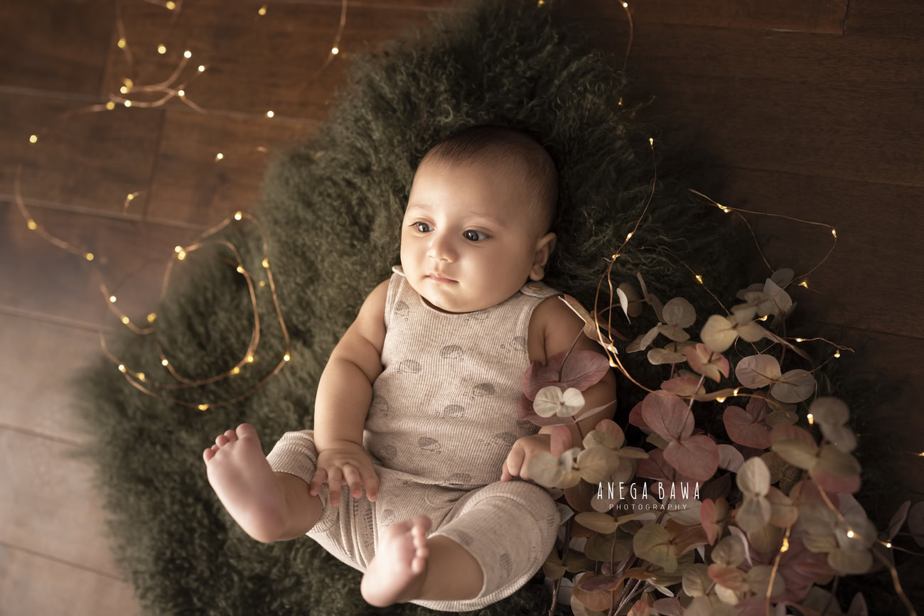 Experience the magic of your baby boy's innocence illuminated by golden lights on a cozy green rug against a warm brown backdrop. Contact Anega Bawa for a memorable photoshoot in Delhi, Gurgaon, or Noida.