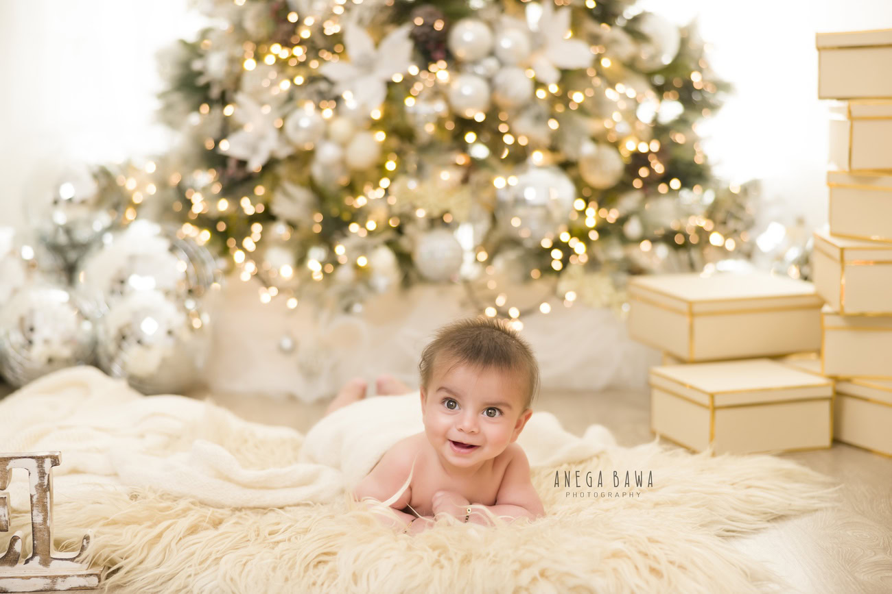 Celebrate your baby boy's milestones with a charming photoshoot featuring an off-white sheet and a cozy beige rug, complemented by twinkling discoballs and adorable gifts, all set against a pristine white backdrop. Contact Anega Bawa for an enchanting baby boy photo session in Delhi, Gurgaon, or Noida.