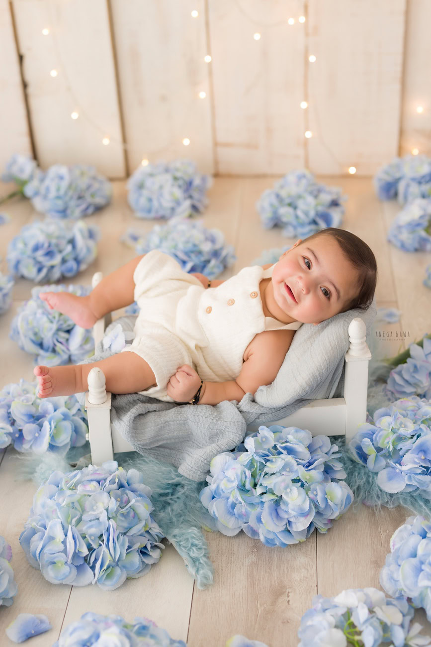 Baby boy photo shoot, 6 or 5 or 3 or 4 months, white baby boy suit, beige backdrop, golden lights, blue flowers. Location: Delhi, Gurgaon, Noida. Photographer: Anega Bawa.