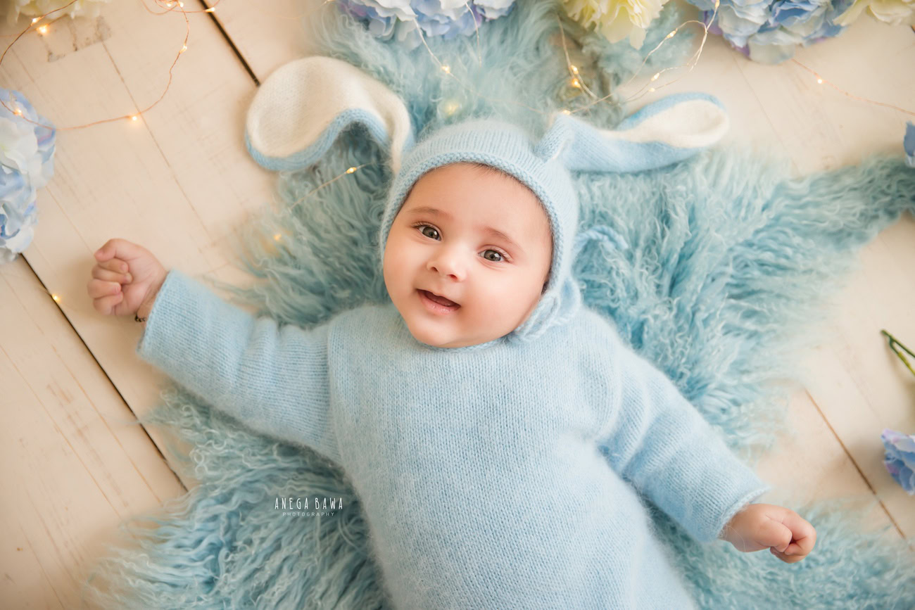 Immerse in the charm of your little prince in a blue baby boysuit, accentuated by a matching cap, on a cozy blue rug, set against a serene beige backdrop. Trust Anega Bawa for captivating baby photography in Delhi, Gurgaon, or Noida.