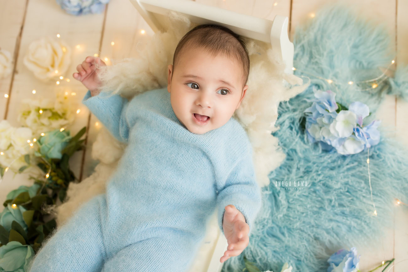 Immerse in the charm of your little prince in a blue baby boysuit, accentuated by a matching cap, on a cozy blue rug, set against a serene beige backdrop. Trust Anega Bawa for captivating baby photography in Delhi, Gurgaon, or Noida.