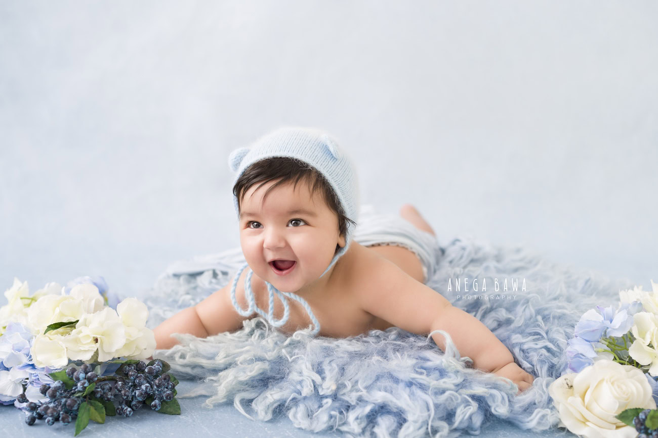 Immerse in the charm of your little one with a heartwarming photoshoot! Your 6, 4, 3, or 5-month-old baby boy adorned in a cute cap lies on a cozy blue rug, surrounded by delicate flowers, set against a serene grey backdrop. Contact Anega Bawa for capturing these precious moments in Delhi, Gurgaon, or Noida.