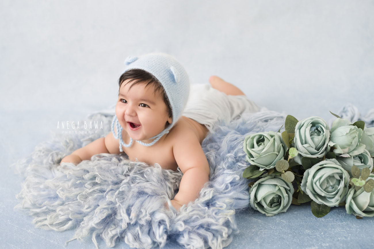 Delight in the charm of your 6, 4, 3, or 5-month-old baby boy, adorned in a cute cap, nestled on a grey rug against a serene blue backdrop with green flowers. Book your session with Anega Bawa for precious memories in Delhi, Gurgaon, or Noida.