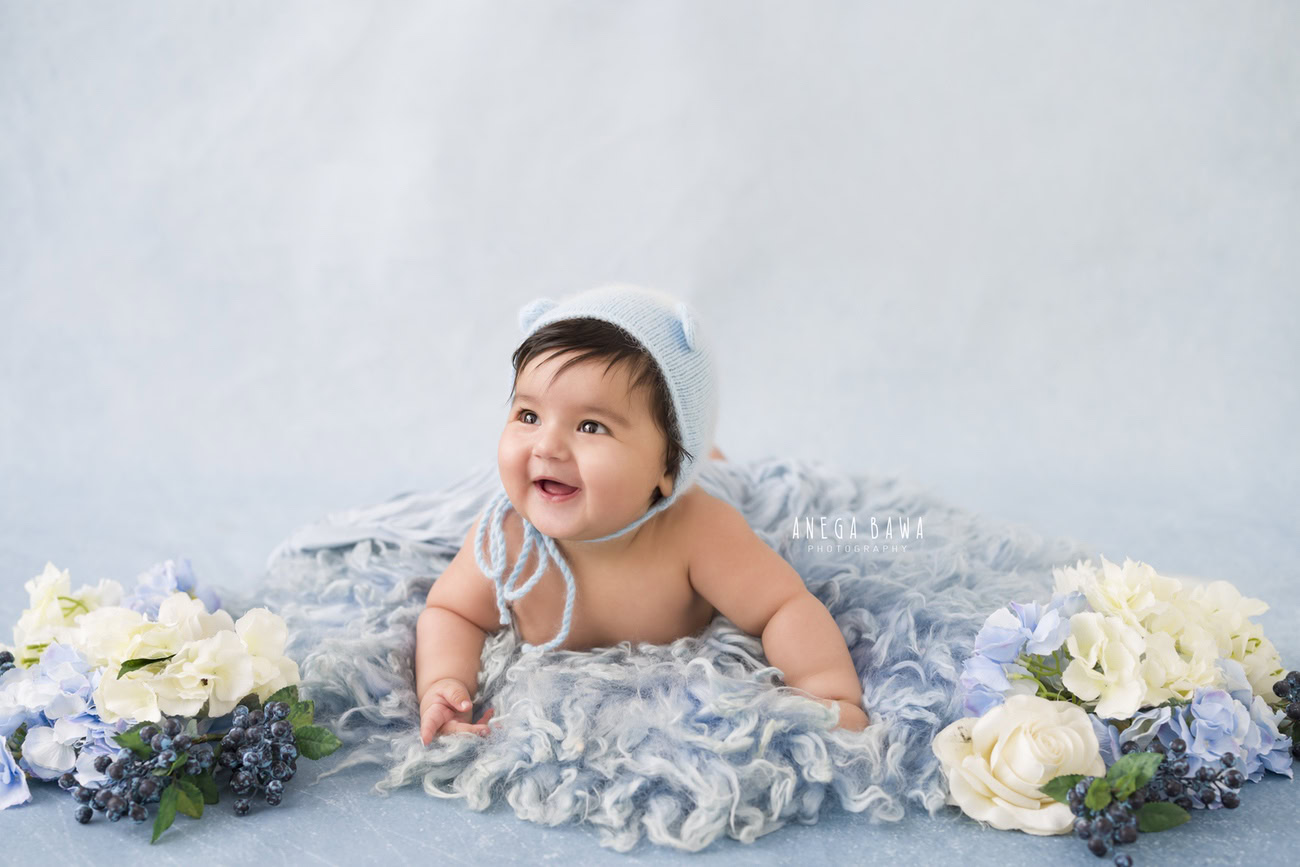 Capture the adorable moments of your 6, 4, 3, or 5-month-old baby boy in a cute cap, set against a grey rug and backdrop adorned with white and blue flowers. Book Anega Bawa for a memorable photoshoot in Delhi, Gurgaon, or Noida.