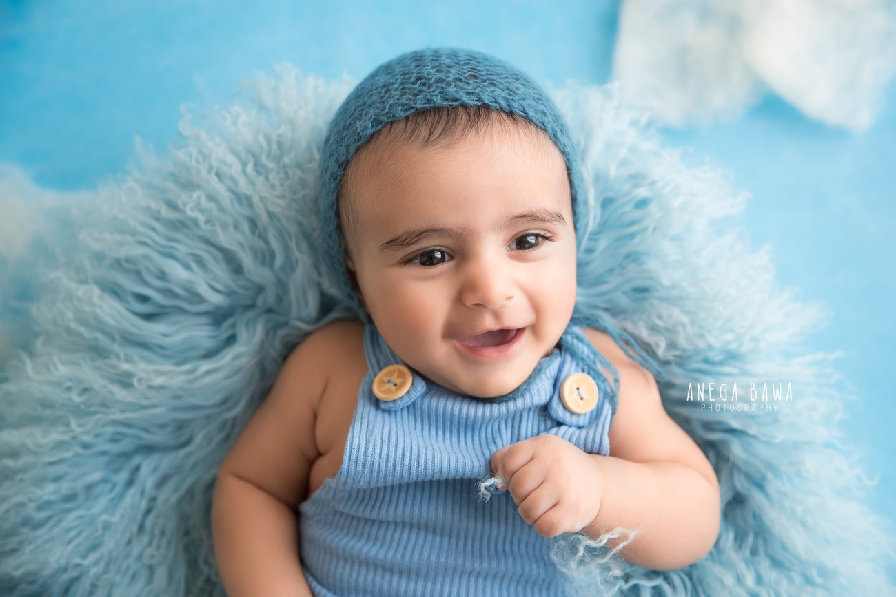 Charming baby boy photography session featuring a delightful 5, 4, 3, 6-month-old adorned with a blue cap, lying on a soft blue rug against a serene blue backdrop. Captured skillfully by Anega Bawa, the esteemed baby photographer in Delhi, Gurgaon, and Noida.