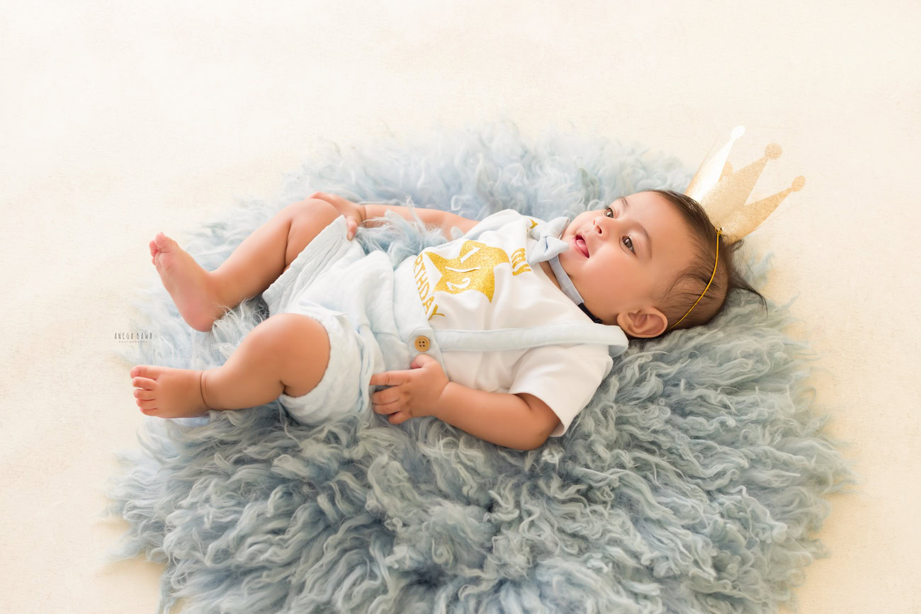 Baby boy photography, 5 or 4 or 3 or 6 months, blue rug, white backdrop, crown on head, lying down pose. Location: Delhi, Gurgaon, Noida. Photographer: Anega Bawa.