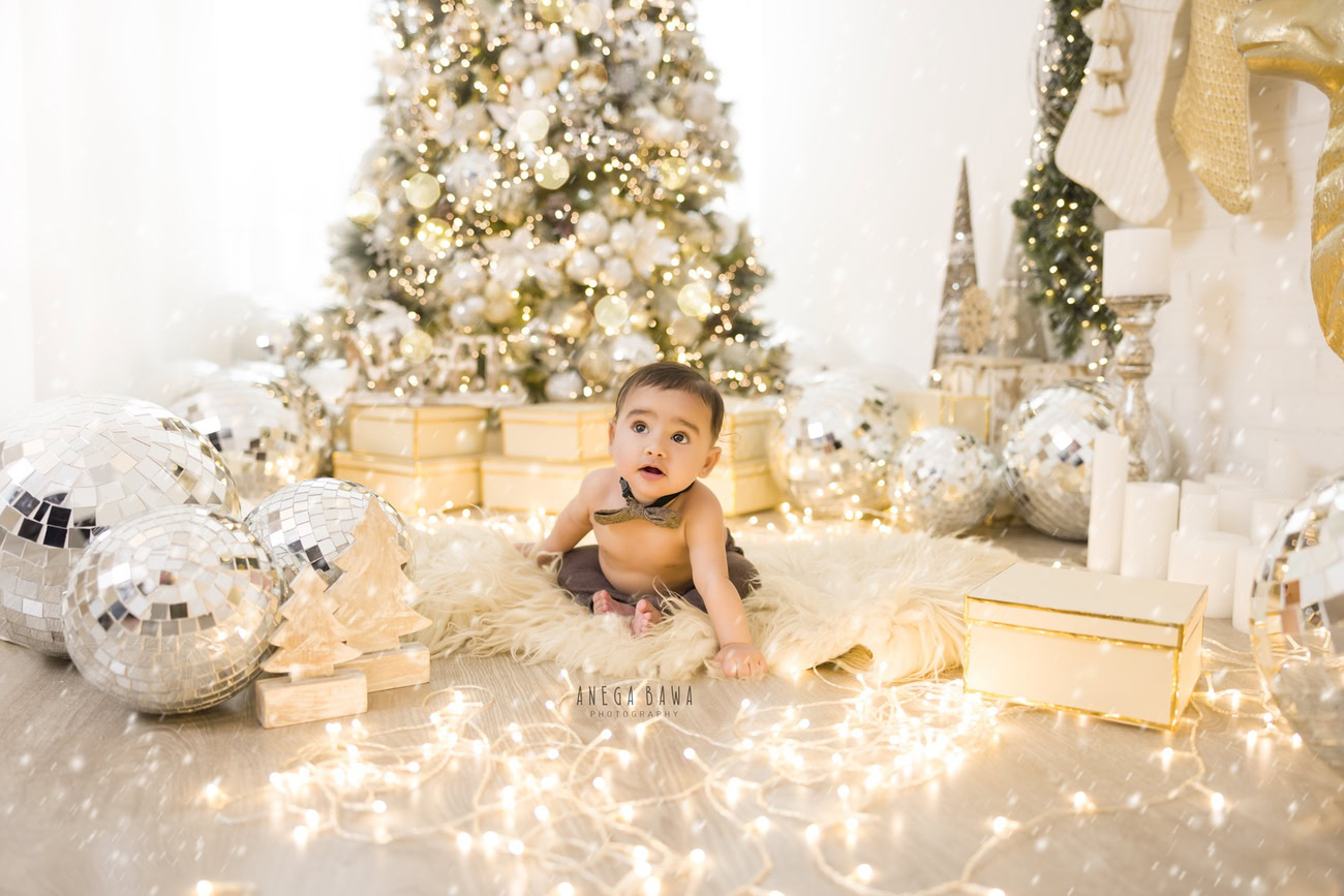 Capture the innocence and joy of your baby boy aged 3, 4, 5, or 6 months with Anega Bawa's enchanting baby boy photography services in Delhi, Gurgaon, or Noida. Adorned with a cute bow, nestled on a cozy beige rug, and surrounded by the festive ambiance of a Christmas tree, golden lights, and disco balls, create magical memories against a pristine white backdrop.