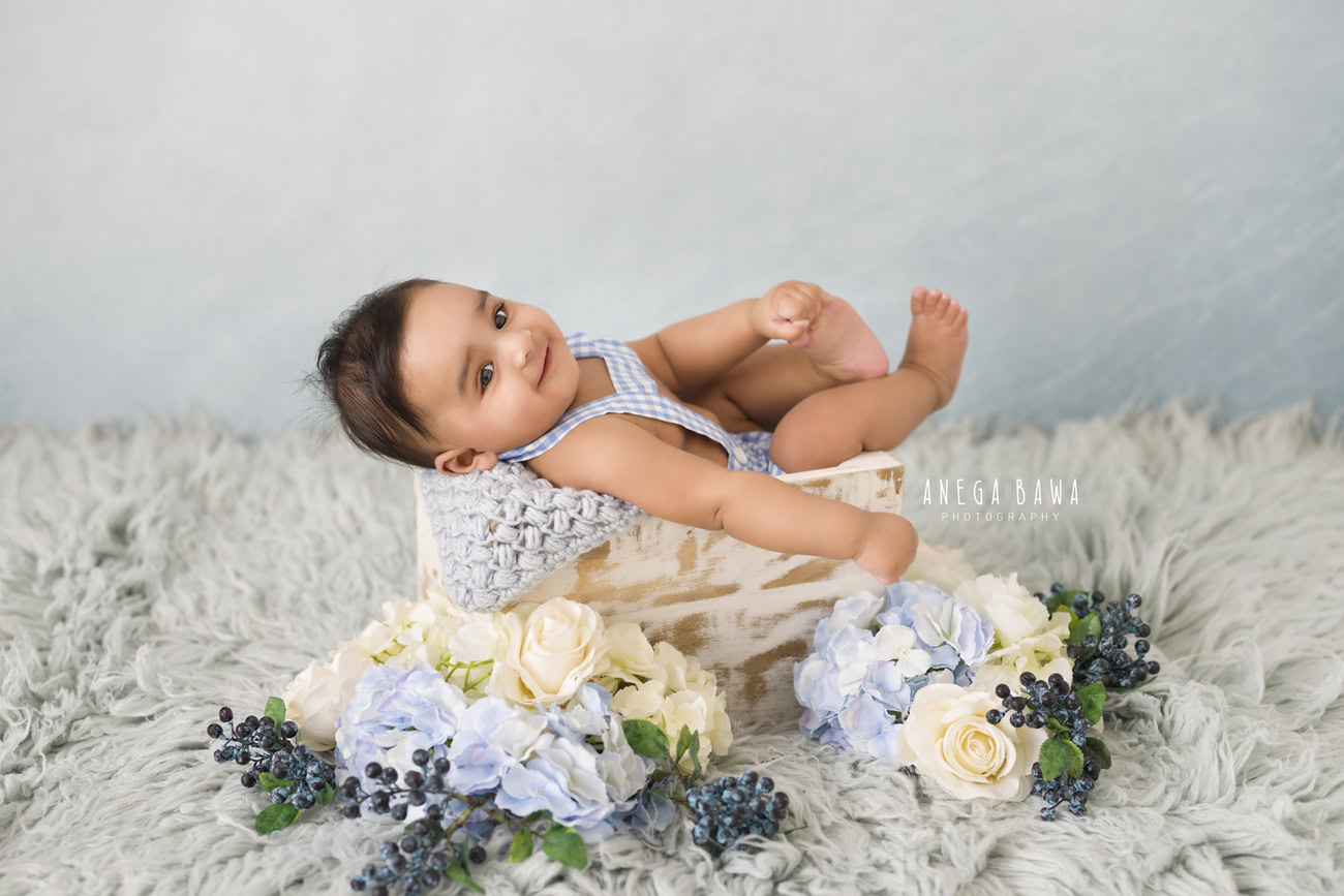 Capture the precious moments of your baby boy aged 3, 4, 5, or 6 months with Anega Bawa's baby boy photography in Delhi, Gurgaon, or Noida. This delightful photoshoot features your little one nestled in a grey basket adorned with blue and white flowers, set against a cozy grey rug backdrop.
