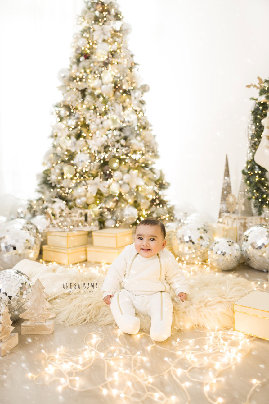 Capture the innocence and charm of your 3, 4, 5, or 6-month-old baby boy in a delightful photoshoot featuring a white baby boysuit against the backdrop of a festive Xmas tree, disco balls, and golden lights. The serene white backdrop adds a touch of elegance to the scene. Contact Anega Bawa for your baby boy's photography session in Delhi, Gurgaon, or Noida.