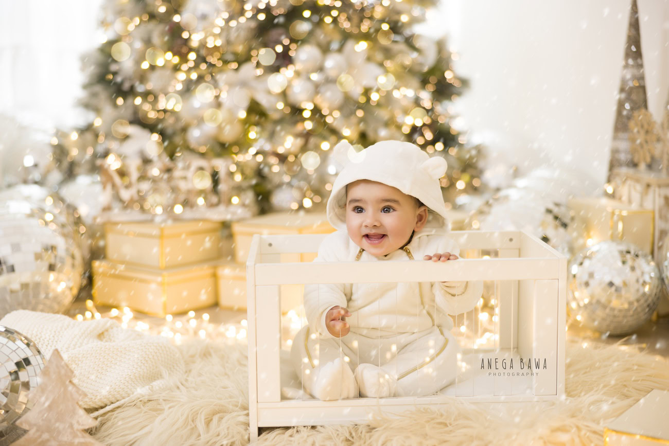 Celebrate the joy of your little one's milestones with our baby boy photography services in Delhi, Gurgaon, or Noida. In this charming photoshoot, your 3, 4, 5, or 6-month-old baby boy wears a cute white baby boysuit and a matching cap, surrounded by the festive ambiance of an Xmas tree and disco balls. The scene is set against a soft beige rug backdrop and a pristine white backdrop. Contact Anega Bawa to capture these precious moments.