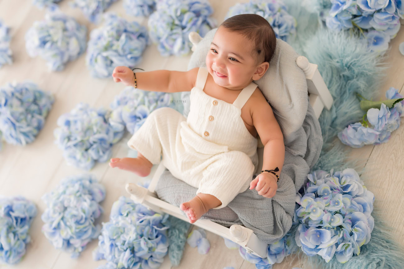 Baby boy photography, 5 or 4 or 3 or 6 months, wooden chair, blue flowers, white baby boy suit, white backdrop. Location: Delhi, Gurgaon, Noida. Photographer: Anega Bawa.