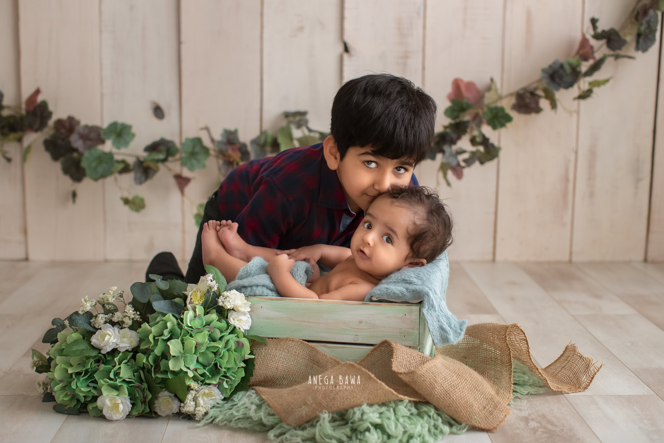 Captivating baby boy photoshoot showcasing the tender moments of siblings, ages 6, 5, 4, 3 months, against a serene beige backdrop, adorned with a gentle blue wrap, nestled in a leafy curve basket. Expertly captured by Anega Bawa, your trusted baby photographer in Delhi, Gurgaon, and Noida.