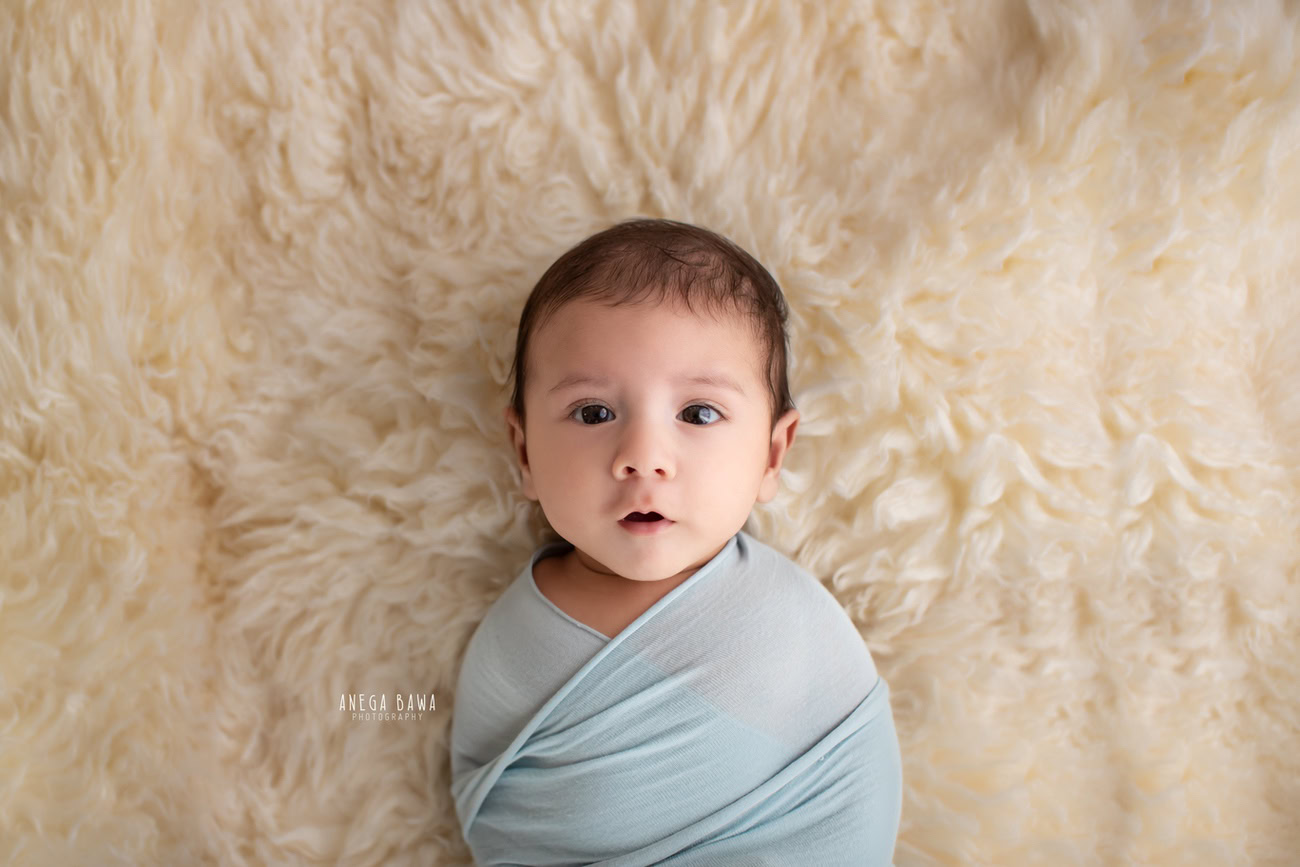 Enchanting baby boy photoshoot featuring a delightful 6, 5, 4, 3-month-old nestled on a cozy beige rug, wrapped in a serene sky blue wrap against a matching beige backdrop. Crafted with expertise by Anega Bawa, the premier baby photographer in Delhi, Gurgaon, and Noida.