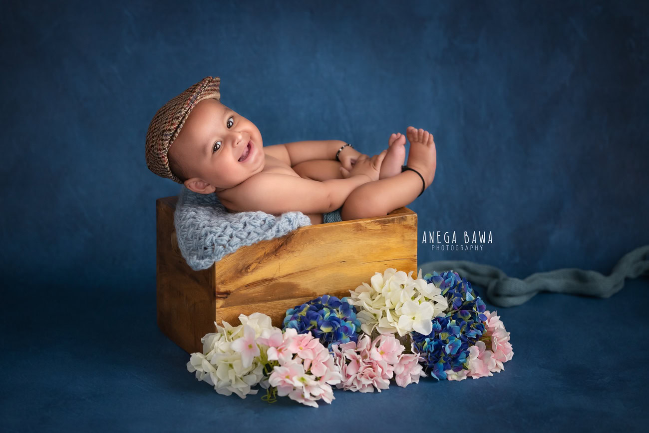 Captivating baby boy photoshoot showcasing the charm of a 6, 5, 4, 3-month-old against a striking blue backdrop, adorned with a cute cap and multicolored flowers. Expertly captured by Anega Bawa, your esteemed baby photographer in Delhi, Gurgaon, and Noida.