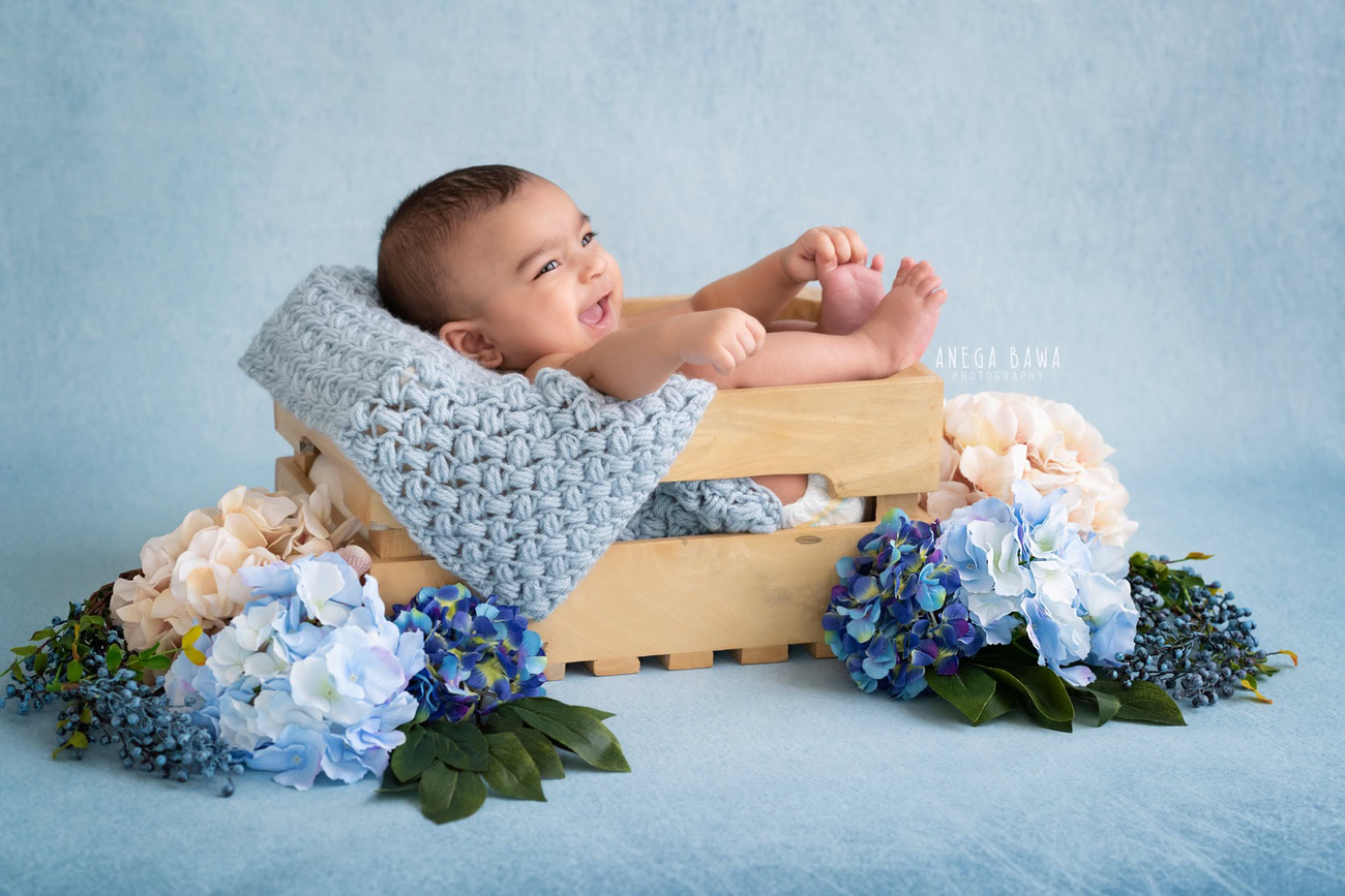 Enchanting baby boy photoshoot featuring a charming 6, 5, 4, 3-month-old against a serene blue backdrop, nestled in a wooden basket adorned with blue and white flowers, showcasing a cute smiling pose. Crafted with expertise by Anega Bawa, the premier baby photographer in Delhi, Gurgaon, and Noida.