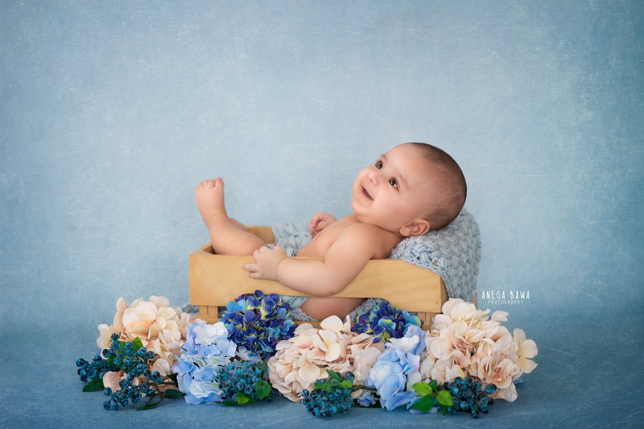 Captivating baby boy photoshoot featuring a delightful 6, 5, 4, 3-month-old against a serene blue backdrop, nestled in a wooden basket adorned with a floral curve, showcasing a cute smiling pose. Crafted with expertise by Anega Bawa, the premier baby photographer in Delhi, Gurgaon, and Noida.