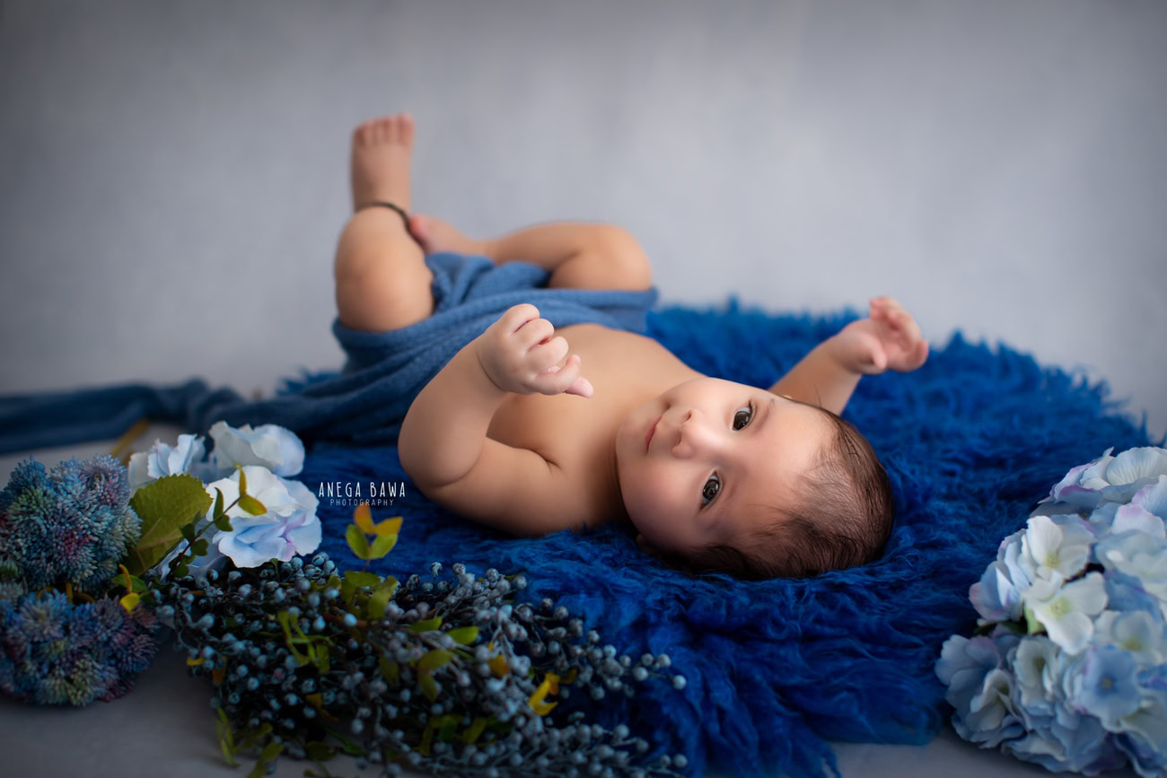 Captivating baby boy photoshoot featuring a delightful 6, 5, 4, 3-month-old on a soft blue rug adorned with blue flowers, wrapped in a serene blue wrap against a stylish grey backdrop. Expertly crafted by Anega Bawa, the premier baby photographer in Delhi, Gurgaon, and Noida.
