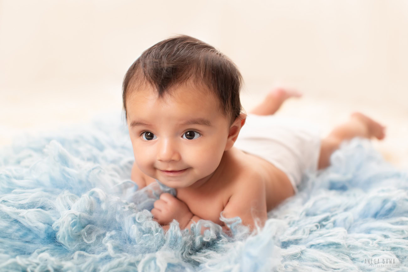 Captivating baby boy photoshoot capturing the adorable 6, 5, 4, 3-month-old on a soft blue rug against a pristine white backdrop, showcasing a cute smiling pose. Expertly crafted by Anega Bawa, the premier baby photographer in Delhi, Gurgaon, and Noida.