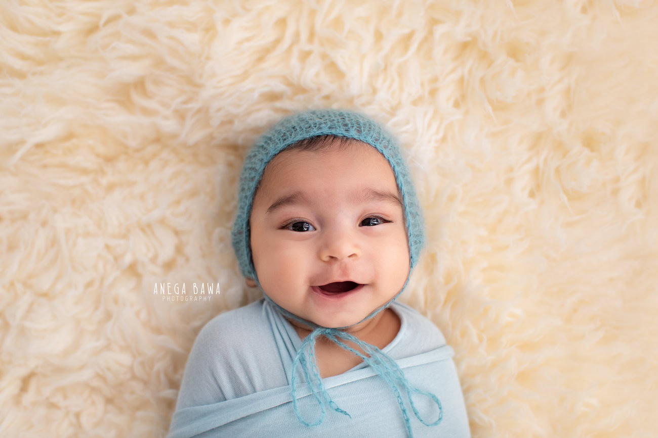 Enchanting baby boy photoshoot presenting the delightful 6, 5, 4, 3-month-old wrapped in a serene blue wrap, adorned with a charming blue cap, resting on a cozy beige rug against a warm beige backdrop. Captured skillfully by Anega Bawa, the renowned baby photographer in Delhi, Gurgaon, and Noida.