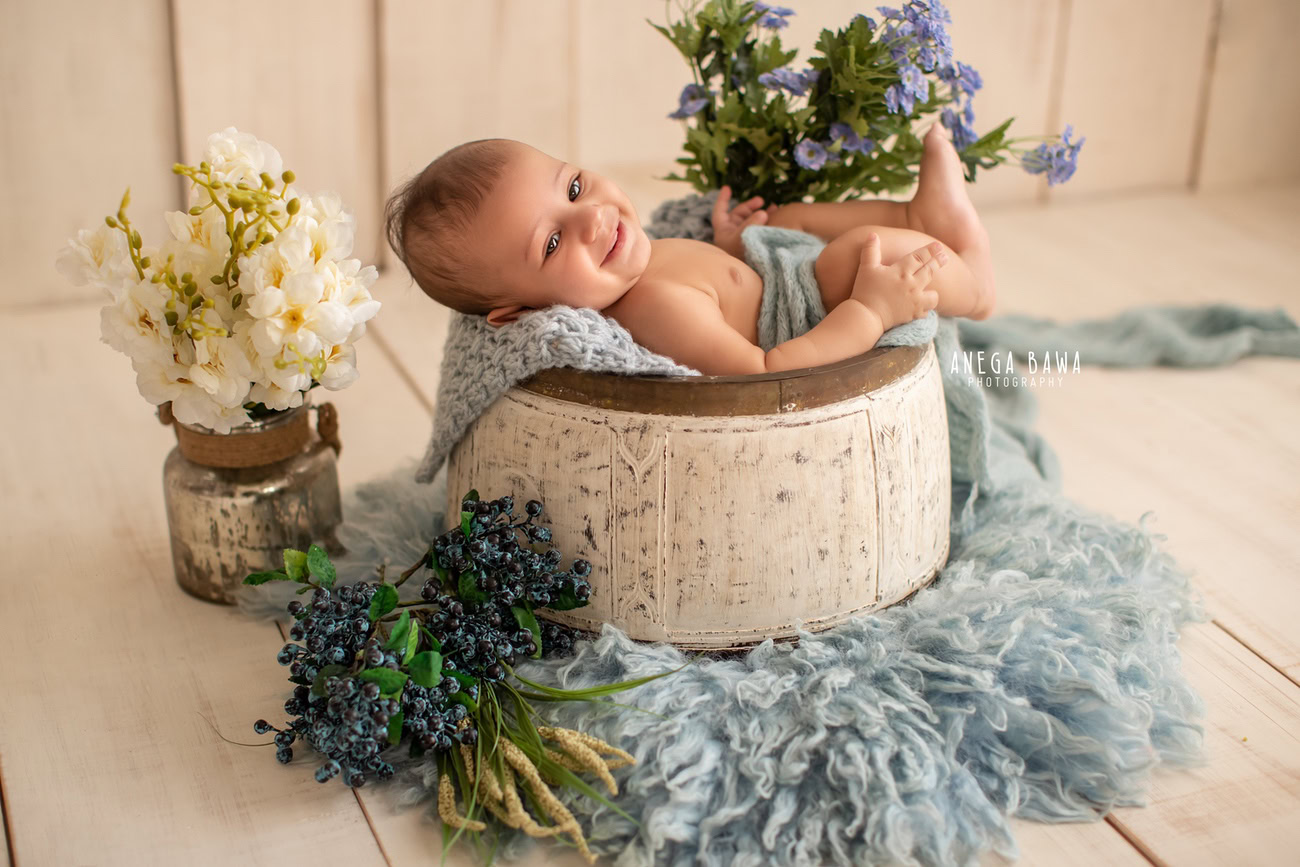 Captivating baby boy photoshoot featuring the enchanting 6, 5, 4, 3-month-old wrapped in a serene blue wrap, lying on a soft blue rug surrounded by a bunch of flowers against a warm beige backdrop. Expertly captured by Anega Bawa, the premier baby photographer in Delhi, Gurgaon, and Noida.