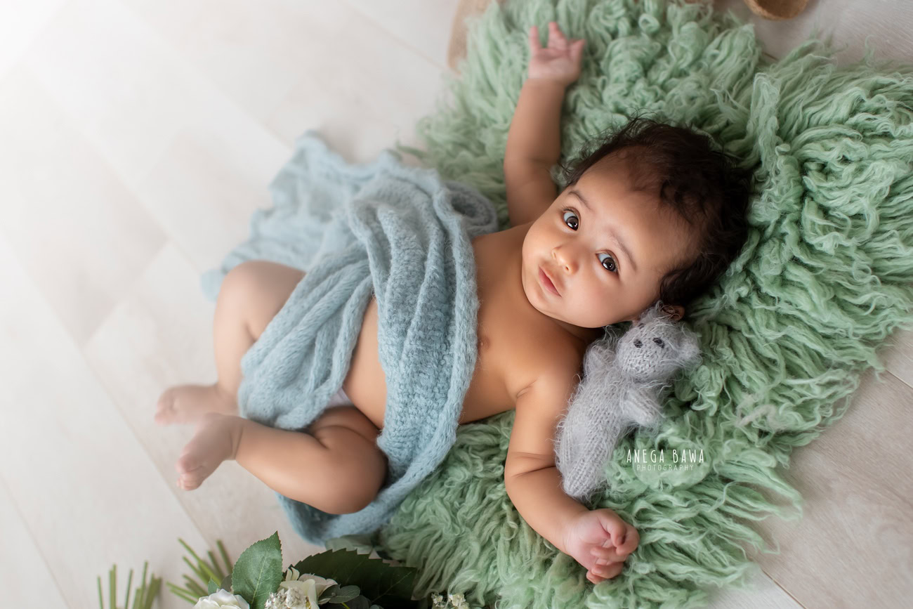 Captivating baby boy photoshoot featuring the charming 6, 5, 4, 3-month-old on a lush green rug, surrounded by a serene blue sheet and soft toy against a warm beige backdrop. Crafted with expertise by Anega Bawa, the premier baby photographer in Delhi, Gurgaon, and Noida.