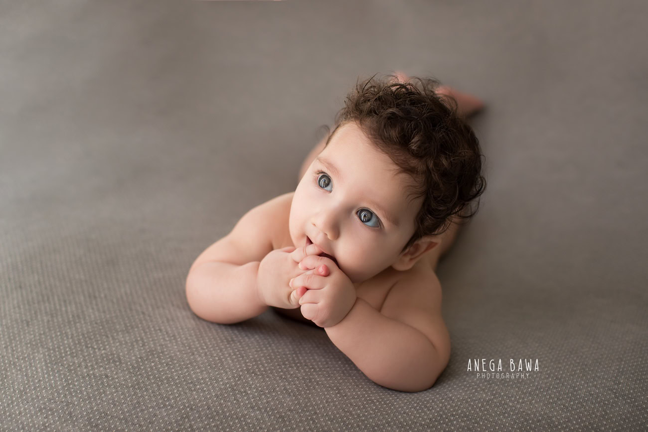 Captivating baby boy photoshoot capturing the adorable 6, 5, 4, 3-month-old in a cute lying pose against a sophisticated grey backdrop. Crafted with expertise by Anega Bawa, the premier baby photographer in Delhi, Gurgaon, and Noida.