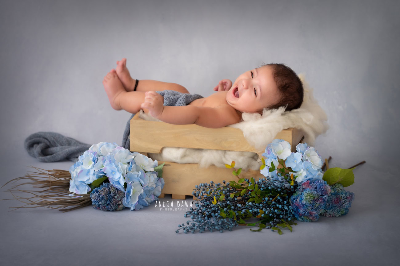Enchanting baby boy photoshoot showcasing the delightful 6, 5, 4, 3-month-old in a smiling pose against a sophisticated grey backdrop, nestled in a wooden cot adorned with blue flowers. Captured with expertise by Anega Bawa, the premier baby photographer in Delhi, Gurgaon, and Noida.