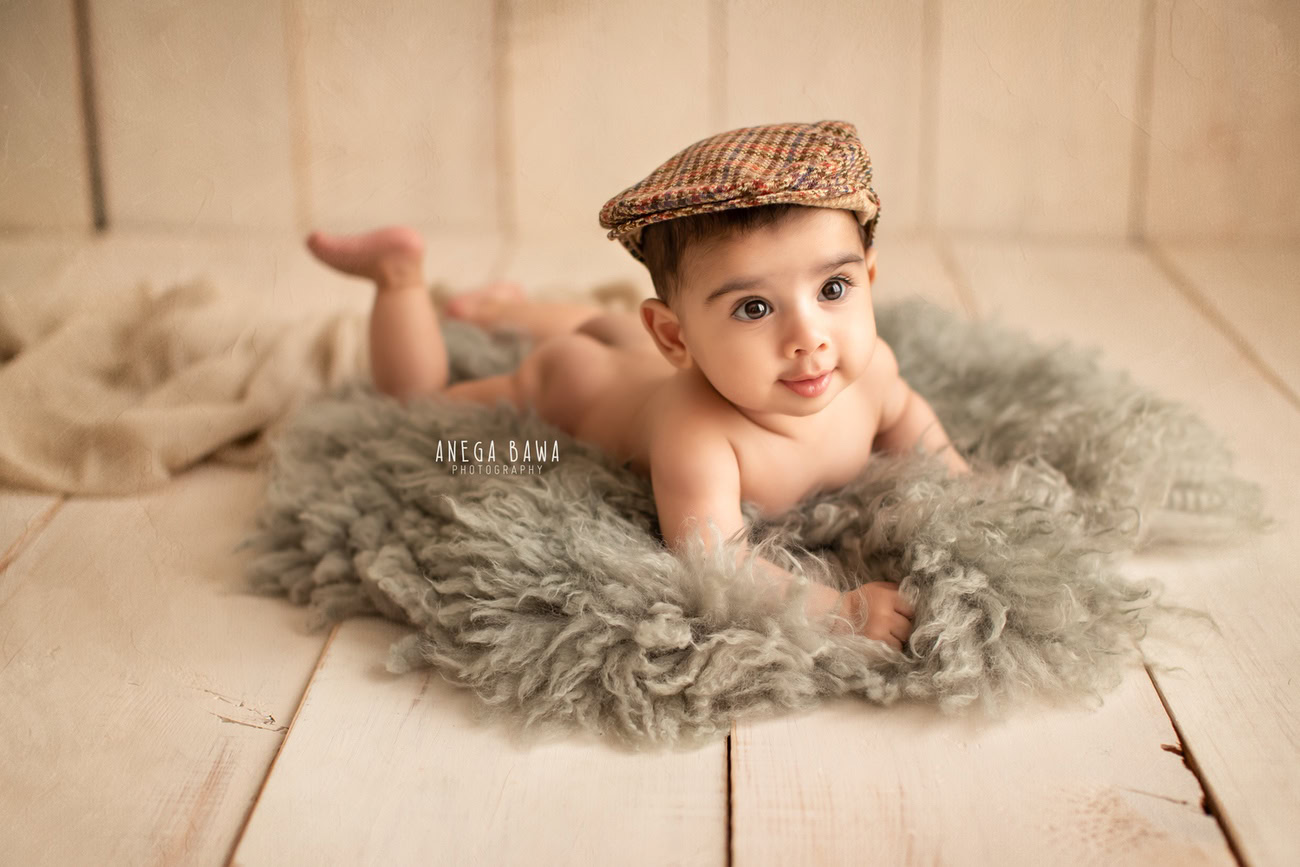 Captivating baby boy photoshoot featuring the adorable 6, 5, 4, 3-month-old adorned with a charming brown cap, resting on a cozy grey rug against a warm beige backdrop. Skillfully crafted by Anega Bawa, the renowned baby photographer in Delhi, Gurgaon, and Noida.