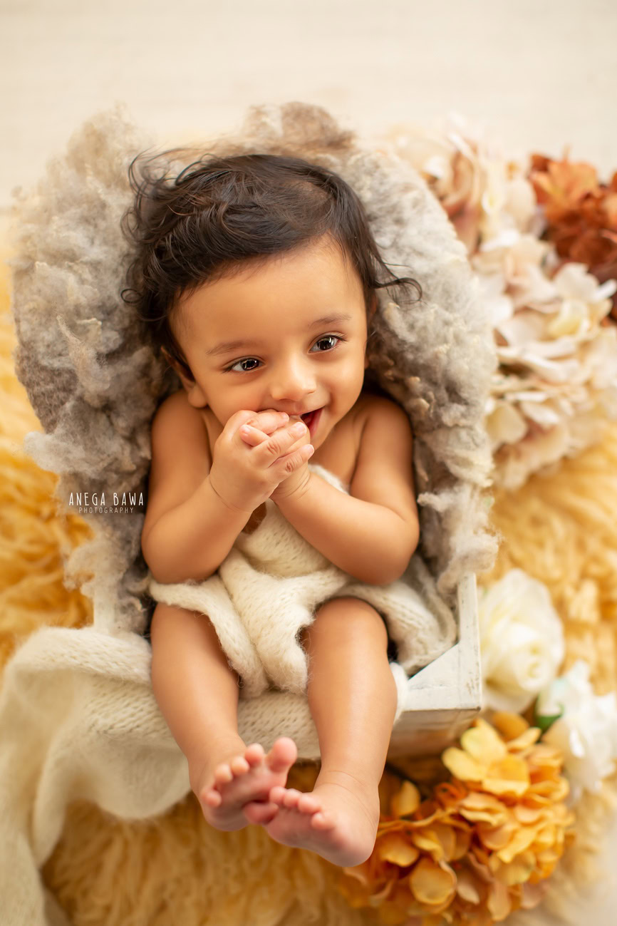 Enchanting baby boy photoshoot capturing the precious 6, 5, 4, 3-month-old in a cute seating pose, surrounded by vibrant yellow flowers on a cozy grey rug against a pristine white backdrop. Skillfully crafted by Anega Bawa, the renowned baby photographer in Delhi, Gurgaon, and Noida.