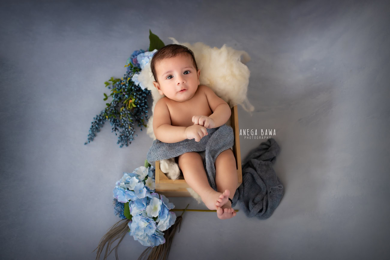 Captivating baby boy photoshoot featuring the charming 6, 5, 4, 3-month-old wrapped in a soft grey wrap against a sophisticated grey backdrop, surrounded by delicate blue flowers in a wooden cot. Skillfully crafted by Anega Bawa, the renowned baby photographer in Delhi, Gurgaon, and Noida.