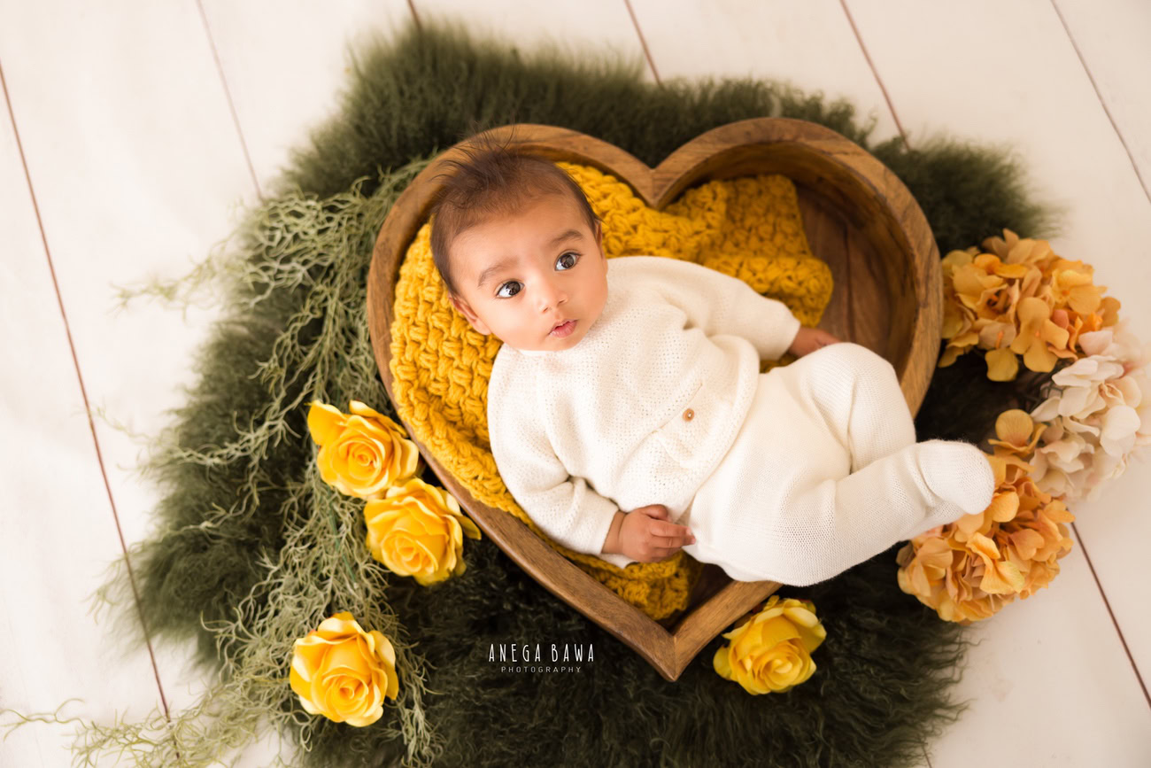 Create precious memories with your baby boy dressed in a white baby boysuit, nestled in a heart-shaped basket surrounded by vibrant yellow flowers, set against a white backdrop. Contact Anega Bawa for a memorable photoshoot in Delhi, Gurgaon, or Noida.