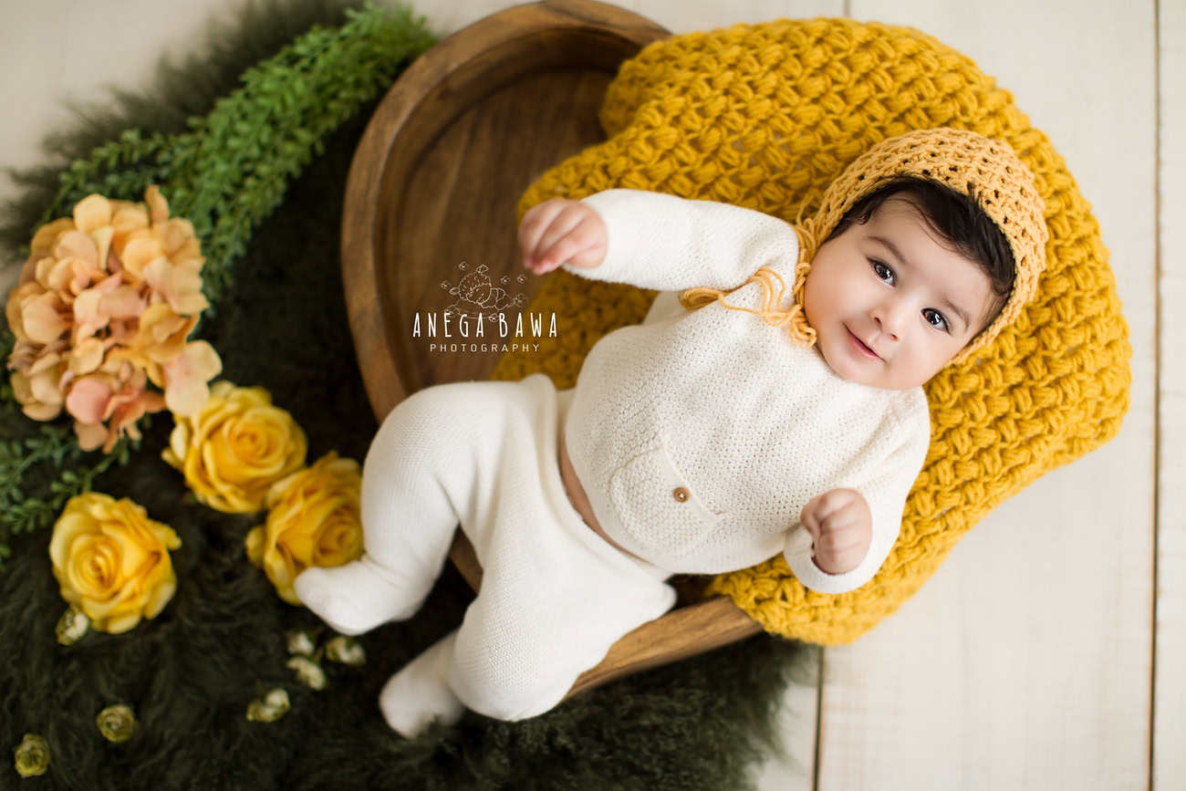 Captivating baby boy photoshoot capturing the innocence of a 6, 5, 4, 3-month-old in a stylish white baby boysuit, complemented by a charming yellow cap, seated on a rustic brown wooden chair. Skillfully orchestrated by Anega Bawa, the esteemed baby photographer in Delhi, Gurgaon, and Noida.