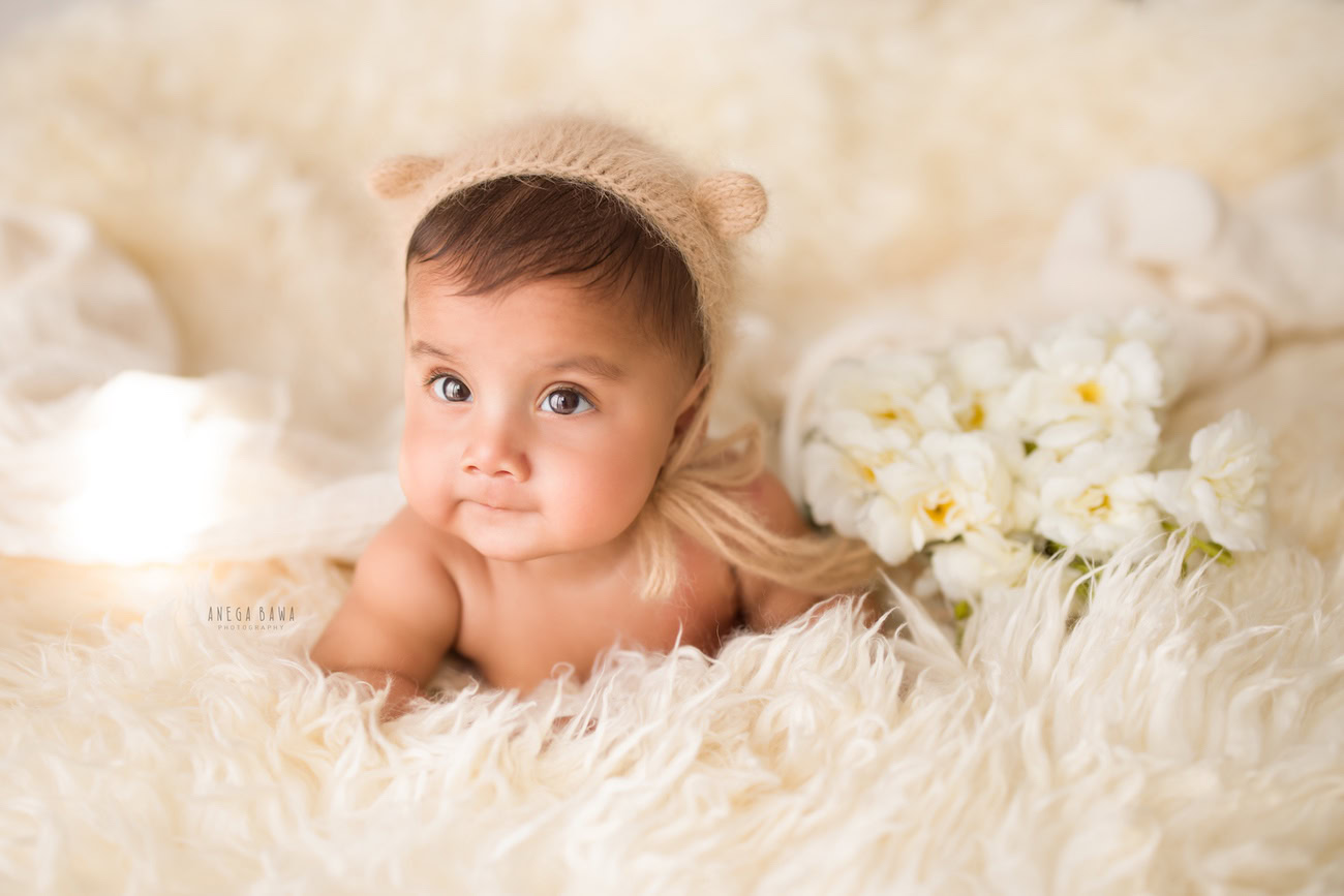 Capture the charm of your baby boy aged 3, 4, 5, or 6 months on a white rug, adorned with a brown cap and surrounded by delicate white flowers. Book your photoshoot with Anega Bawa in Delhi, Gurgaon, or Noida.