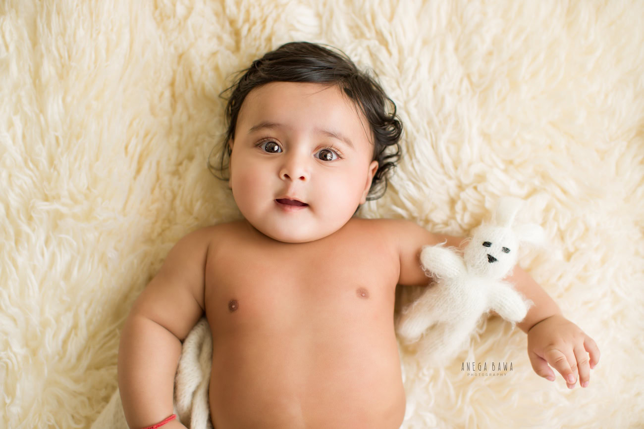 Captivating baby boy photoshoot showcasing the innocence of a 6, 5, 4, 3-month-old on a soft white rug, accompanied by a cuddly white soft toy against a warm beige backdrop. Skillfully captured by Anega Bawa, the esteemed baby photographer in Delhi, Gurgaon, and Noida.