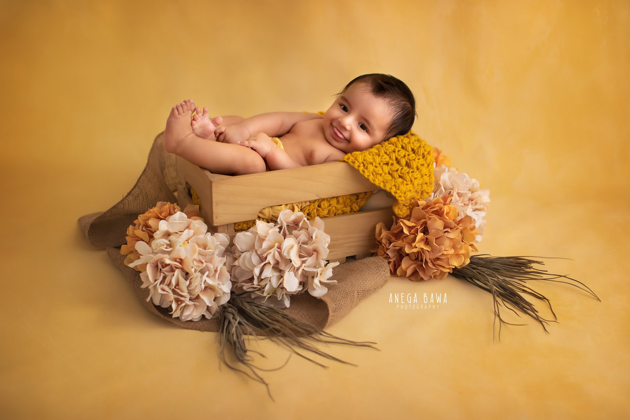 Captivating baby boy photoshoot featuring the delightful 6, 5, 4, 3-month-old nestled in a rustic wooden basket against a cheerful yellow backdrop, adorned with vibrant orange and peach flowers. Skillfully crafted by Anega Bawa, the premier baby photographer in Delhi, Gurgaon, and Noida.