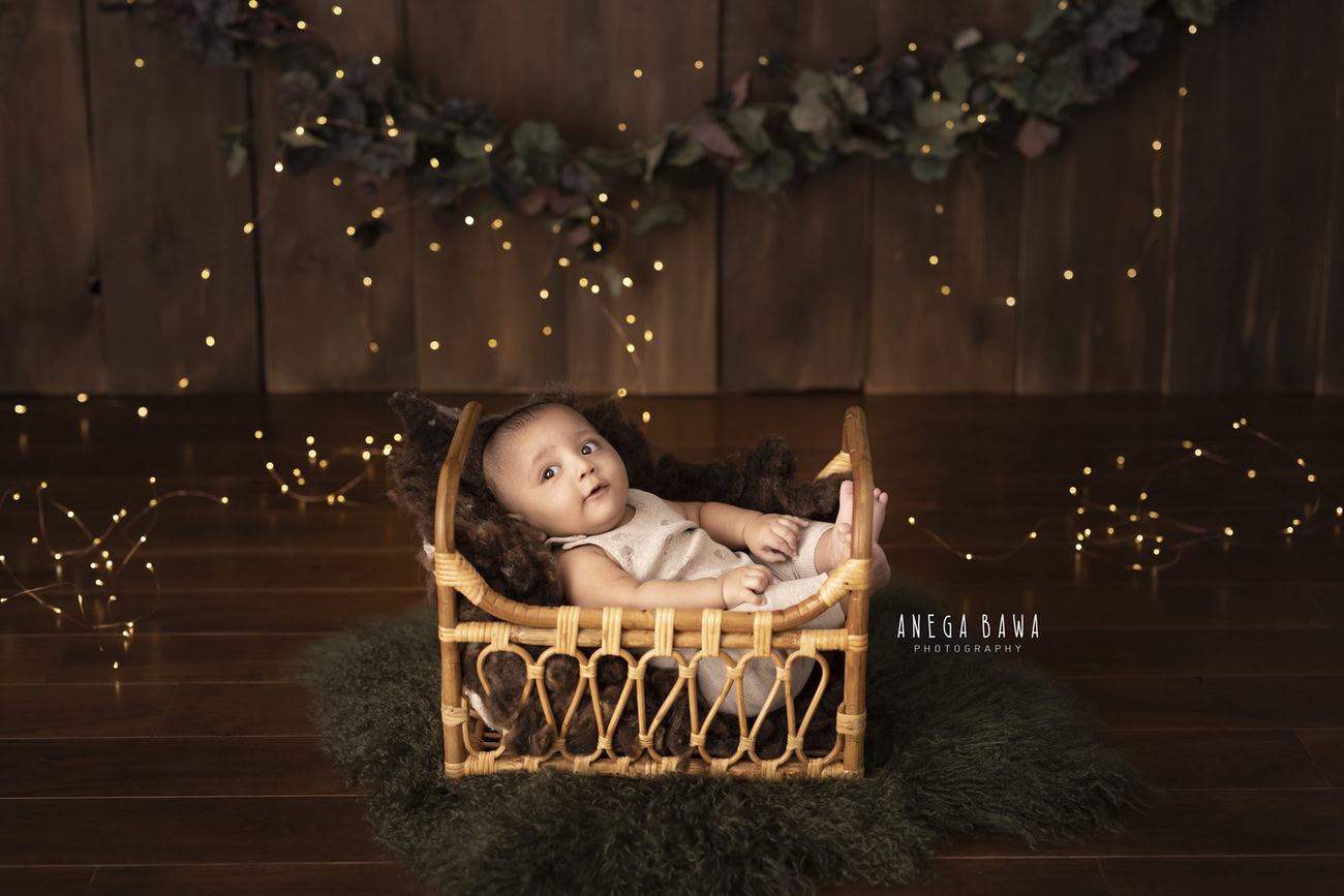 Capture the warmth and charm of your baby boy's early months with a cozy wooden cot, illuminated by soft golden-brown lights, against a warm brown backdrop. Book your photoshoot with Anega Bawa in Delhi, Gurgaon, or Noida.