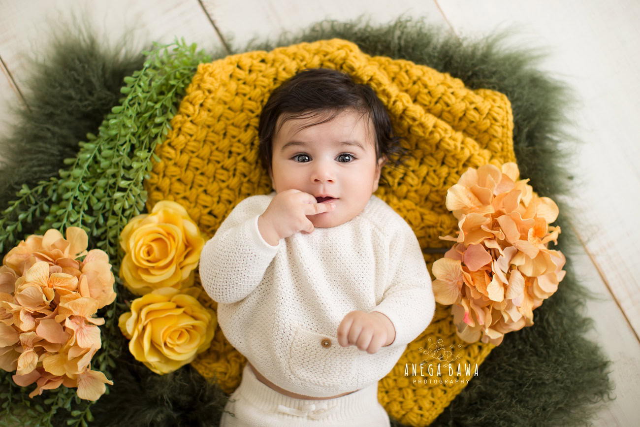 Captivating baby boy photoshoot featuring the charming 6, 5, 4, 3-month-old adorned in a stylish white baby boysuit on a vibrant yellow and green rug against a pristine white backdrop. Skillfully captured by Anega Bawa, the esteemed baby photographer in Delhi, Gurgaon, and Noida.