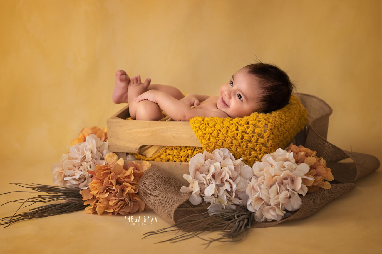 Enchanting baby boy photoshoot capturing the precious 6, 5, 4, 3-month-old against a cheerful yellow backdrop, nestled in a rustic wooden basket surrounded by vibrant orange and peach flowers and brown bushes. Skillfully crafted by Anega Bawa, the premier baby photographer in Delhi, Gurgaon, and Noida.